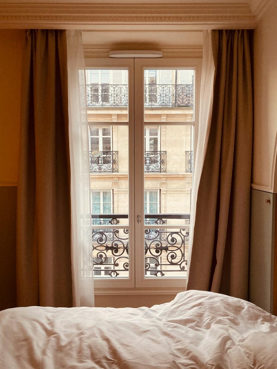 Cama junto a un balcón alto con cortinas largas y, enfrente, balcones antiguos de hierro